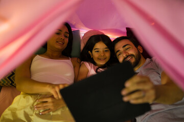 Smiling family enjoying a night in, watching movies under a colorful blanket fort with soft lighting creating a warm ambiance