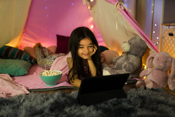 Happy young girl has a cozy movie night in a homemade tent, with plush toys and twinkling lights as company