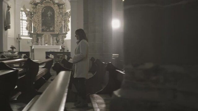 Mid adult woman walking through church aisle, kneeling down and crossing herself side view tracking shot. Christian female parishioner praying slow motion