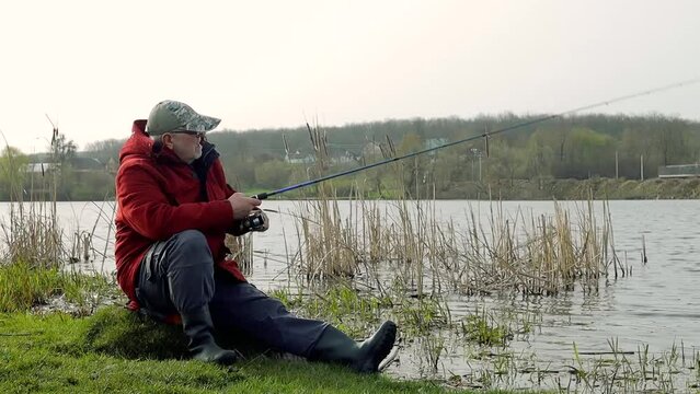Mature adult senior angler fisherman sitting on the rock near river with fishing rod tackle in search of a catch, wear boots and vest, casts his line into water, reel hand rotation, nature
