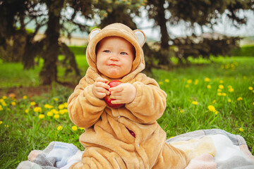 Cute baby girl in plush suit with red apple in hands sitting on blanket in green grass.