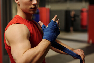 Cropped shot of young male athlete in red vest standing in front of camera in gym and wrapping hand...