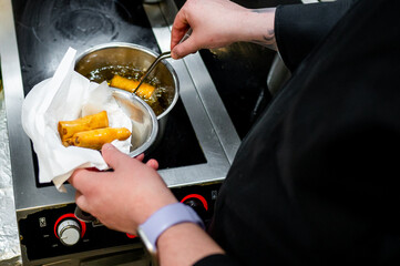 A chef carefully fries spring rolls in a stainless steel pan, using tongs to transfer them to a paper towel for oil absorption