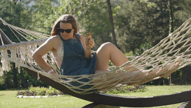 A young woman with ice cream in a hammock sits on the edge and falls backward with it, she screams. A funny incident. Slow motion.