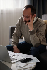 Young pensive or troubled man sitting by table with financial bills and looking at laptop screen...