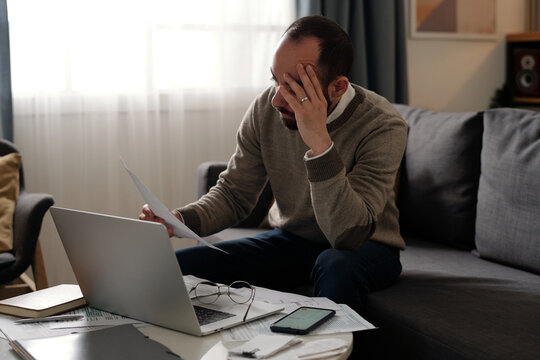 Young troubled man touching his head and looking through financial bill while sitting on couch in front of table with laptop and papers