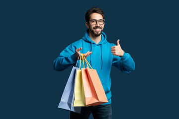 A man wearing a blue hoodie is standing while holding shopping bags in his hands. He is smiling and...