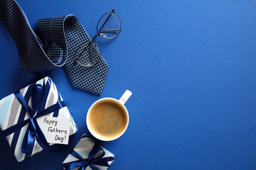 Happy Fathers Day gift box, necktie, glasses, coffee cup on dark blue table, top view.