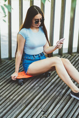 Caucasian woman 20s in trendy apparel spending time with smartphone and skateboard outdoors, attractive stylish hipster girl in sunglasses reading news in social network using cellular phone