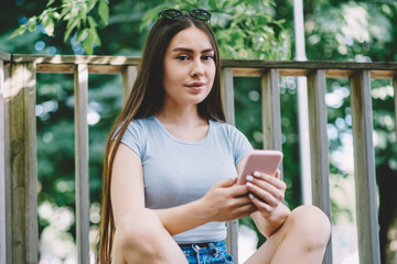 Portrait of young positive woman looking at camera while sitting on skateboard with mobile phone in hand, brunette hipster girl holding modern smartphone and chatting using application outdoors
