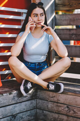 Good looking young female skater talking on smartphone while sitting on urban setting outdoors, trendy teenage woman 20 years old calling via new modern mobile phone and good connection in roaming