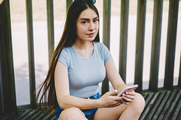 Portrait of young female skater looking at camera and blogging via new telephone in hand, charming hipster girl using modern mobile phone and sending text message while browsing internet outdoors