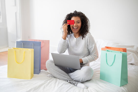 A Hispanic woman wearing casual clothes is sitting on a bed surrounded by colorful shopping bags and a laptop, cover one of her eyes with credit card