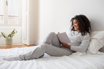 A Hispanic woman in casual clothing sitting on a neatly made bed, engrossed in writing in diary....