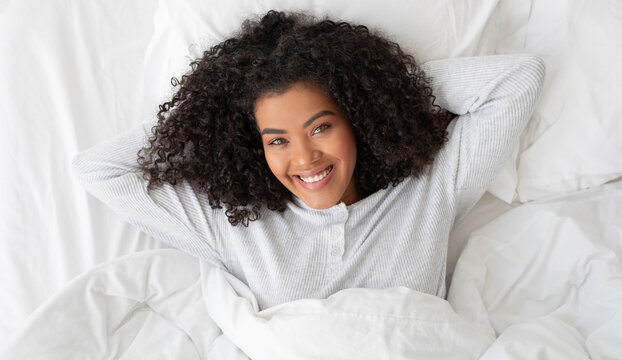 A young Hispanic woman with curly hair is lying on her back in a white bed, resting her head on a pillow. She looks into the camera with a bright, engaging smile