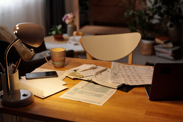 Part of wooden table with lamp illuminating open notebook with envelope containing dollars, unpaid financial bills, cup of tea and gadgets