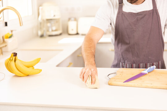 A man is cleaning a cutting board with a cloth. He is wearing an apron and has a knife on the cutting board