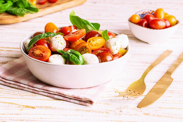 Fresh juicy homemade caprese salad with baby mozzarella cheese, cherry tomatoes, basil leaves and olive oil on white wooden table with colden cutlery set and napkin. Traditional Italian cuisine.