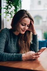 Smiling successful female student watching funny video vlog on website blogger via modern cellular phone while resting at cozy cafeteria, concept of millennial lifestyle and advanced technology