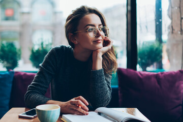 Dreaming beautiful hipster girl looking away while resting at cafeteria and writing romantic essay,...