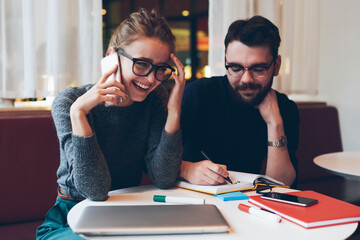 Cheerful laughing hipster girl in eyeglasses spending time with male bearded man while talking via...