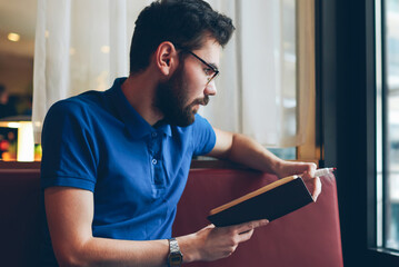 Young pensive hipster guy spending time at university cafeteria for studying and writing plan of...
