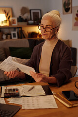 Aged serious woman with white hair sitting by table in home environment and looking through paid...