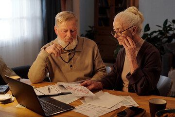 Troubled senior couple sitting by table with financial bills and laptop and looking at unpaid...