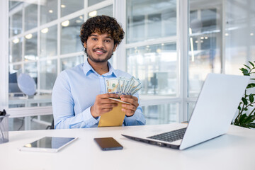 Young businessman in a modern office, smiling as he holds an envelope full of cash. This image...