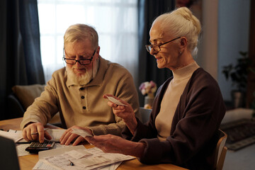 Senior husband and wife checking sums of paid financial bills while man with white beard using calculator and woman looking at receipt