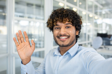 A cheerful young man with curly hair greeting enthusiastically in a modern office environment. He's...