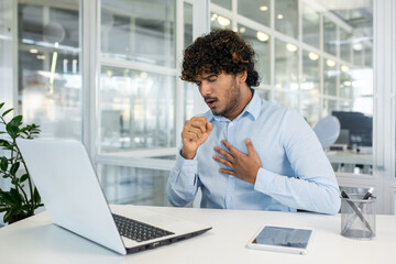 A young man in a light blue shirt feels a sudden cough while working on his laptop in a modern office setting. Concern and discomfort are evident as he addresses his health at work.