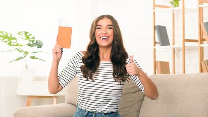 A delighted young woman stands in a sunny living room, waving her passport and giving a thumbs up,...