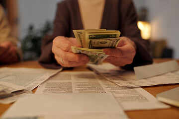 Hands of senior female owner of flat counting dollar banknotes over financial paper documents while sitting in front of camera