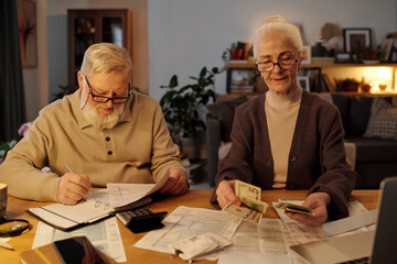 Aged man with financial bills in hand making notes on page of open notebook while sitting next to his wife counting dollar banknotes