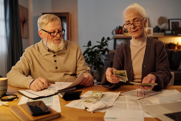 Bearded senior man with stack of financial paper documents in hand looking at his wife counting dollars while sitting by table next to her