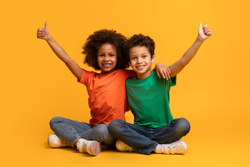 Two African American children are sitting on the floor with their arms stretched up in the air, showing thumb ups. They look joyful and energetic as they play together. - Powered by Adobe