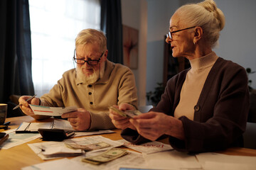 Elderly man with white beard and hair looking through posted financial bill while sitting by table next to his wife counting dollar banknotes