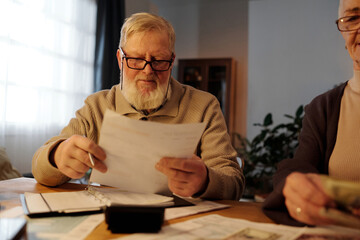 Serious mature man with white beard holding financial document with housing payment sums and...