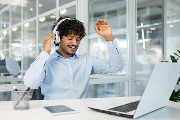 A cheerful young man with curly hair, wearing headphones, happily listens to music while dancing at his office desk, surrounded by modern technology.