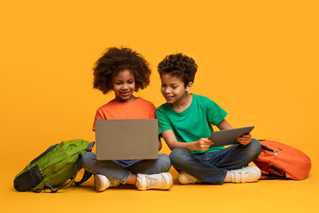 Two young African American children are sitting cross-legged on the floor, engrossed in using a laptop and digital tablet together.