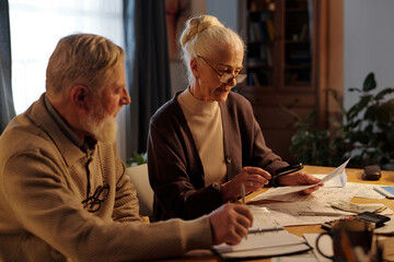 Aged woman in casualwear and eyeglasses holding magnifying glass over financial bill and reading it while sitting next to her husband