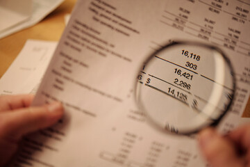 Focus on magnifying glass held by senior person holding financial bill with unpaid sums and...