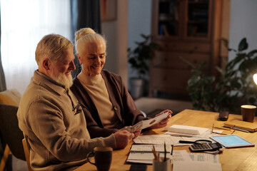 Smiling and surprised elderly husband and wife reading document sent by post and expressing gladness about winning sum of money