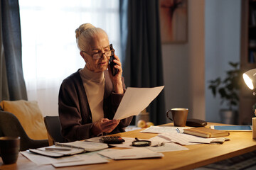 Troubled elderly woman with mobile phone by ear looking at financial paper document while checking information in payment bill