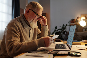 Side view of serious elderly man in eyeglasses checking payment information on receipts while...