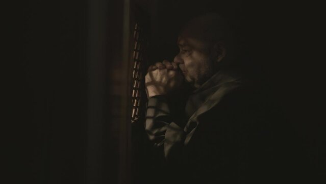 Mid adult monk sitting in confessional booth and praying for forgiveness. Caucasian bald Christian clergyman listening to confession, crossing and speaking