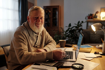 Smiling senior man with white beard looking at camera while sitting by table with laptop and...