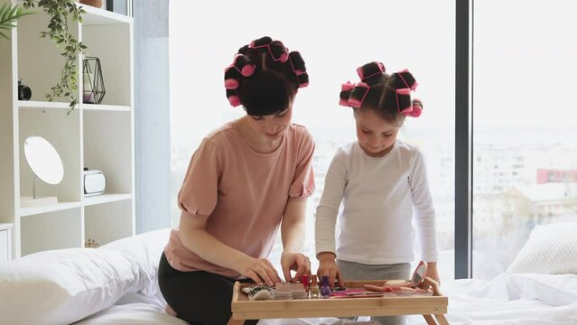 Caucasian family mother and daughter perform cosmetic procedures at the weekend while sitting on the bed. Young woman and cute girl take eye patches from cosmetic table.