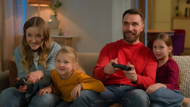 Caucasian family mom dad playing online cyber video game home weekend fun gaming addiction happy joyful children kids little girls daughters and parents play console joysticks celebrate win hive five
